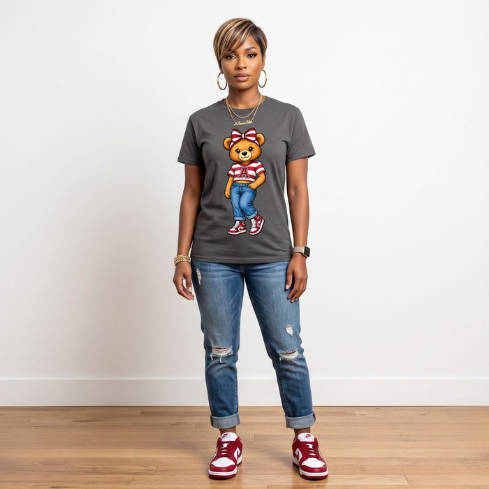 A woman modeling a dark gray graphic t-shirt featuring a cartoon teddy bear wearing a red and white striped shirt and blue jeans, paired with distressed blue jeans and red sneakers, standing against a white wall and light wood floor.