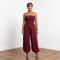 A woman modeling a strapless burgundy jumpsuit with a gathered waist and wide, balloon-style legs, standing against a white wall on a wood floor.