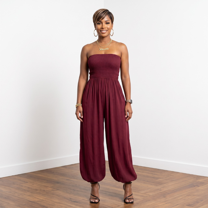 A woman modeling a strapless burgundy jumpsuit with a gathered waist and wide, balloon-style legs, standing against a white wall on a wood floor.
