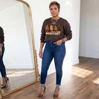 African American woman modeling House of Charlotte HERSTORY graphic sweatshirt styled with dark denim skinny jeans and chocolate brown pumps in a modern room with white walls, hardwood floors, and gold framed mirror.
