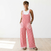 A woman modeling a sleeveless red and white striped wide-leg jumpsuit with front patch pockets, worn over a white tank top, in a bright studio setting with a white wall and light wood floor.