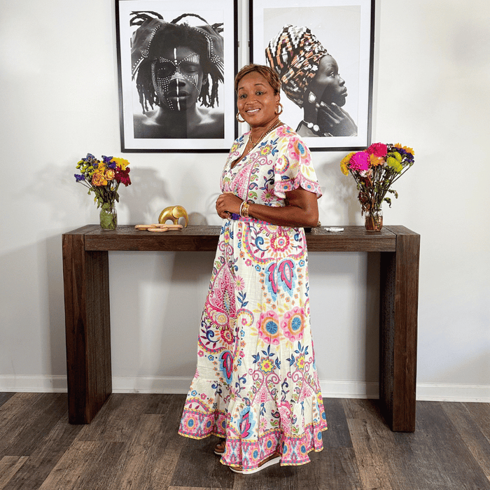 Woman in a colorful dress standing in a room with a wooden console table, vases, and framed art on the wall.