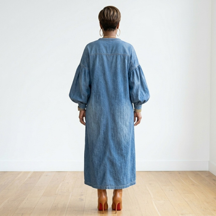 Back view of the Lena Denim Dress on a model, showing its relaxed silhouette and long lantern sleeves in a white room with hardwood flooring.