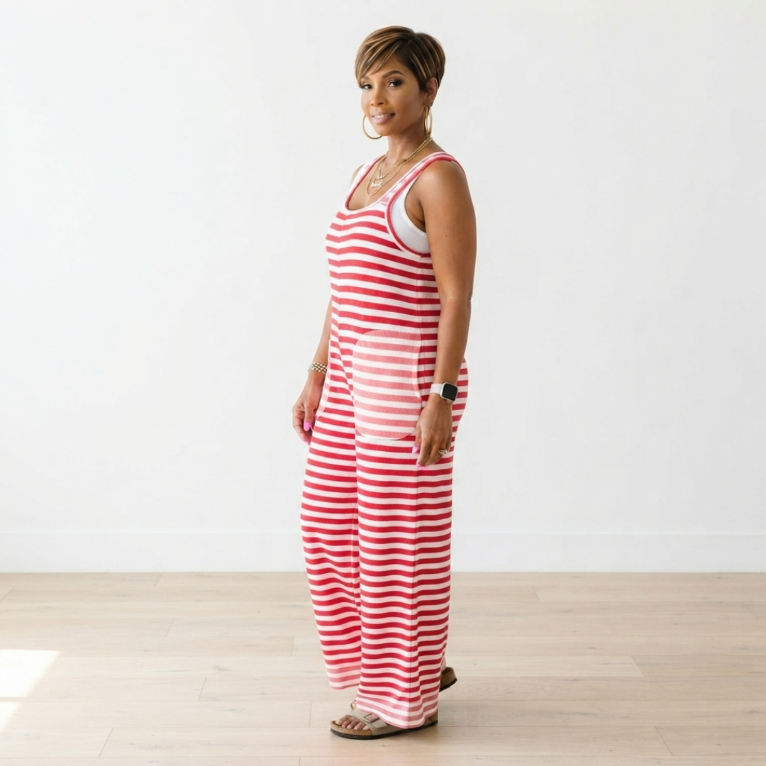 A woman modeling a red and white striped sleeveless jumpsuit with a front pocket, standing in a bright room with a white wall and light wood floor.