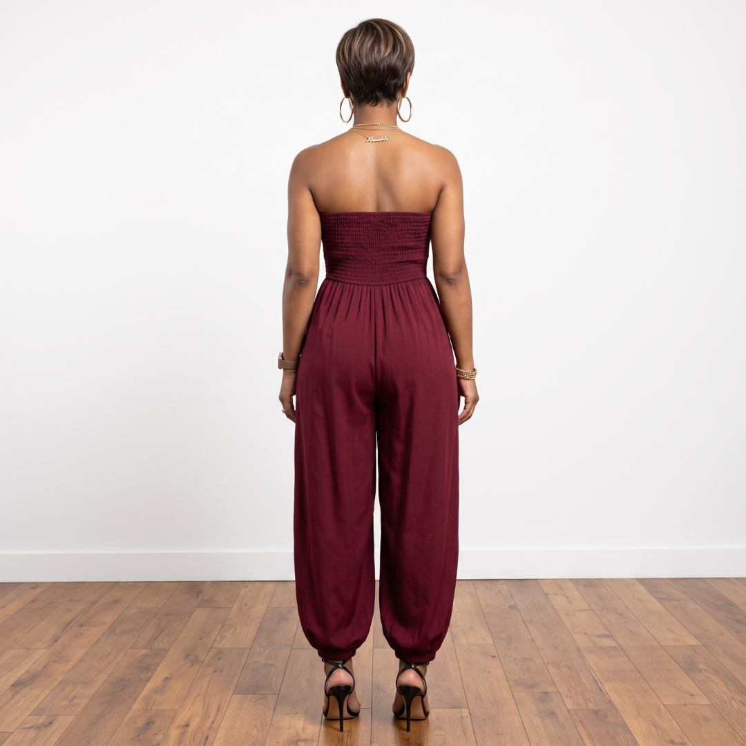 A woman viewed from behind wearing a strapless burgundy jumpsuit with a ruched bodice and wide-leg harem-style pants, standing on a wood floor against a plain white wall.