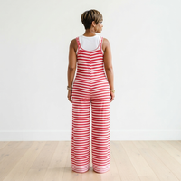 A woman viewed from behind wearing a red and white striped sleeveless jumpsuit over a white tank top, standing on a light wood floor against a white wall.