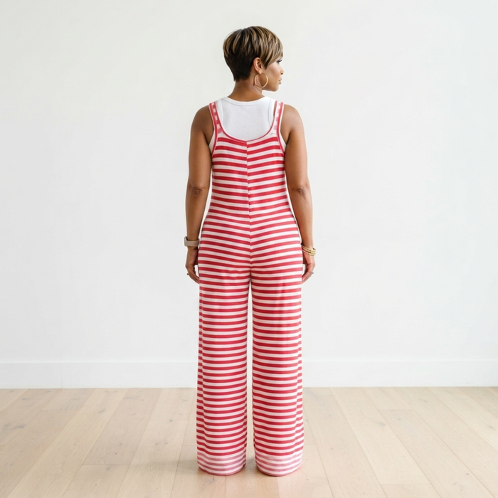 A woman viewed from behind wearing a red and white striped sleeveless jumpsuit over a white tank top, standing on a light wood floor against a white wall.