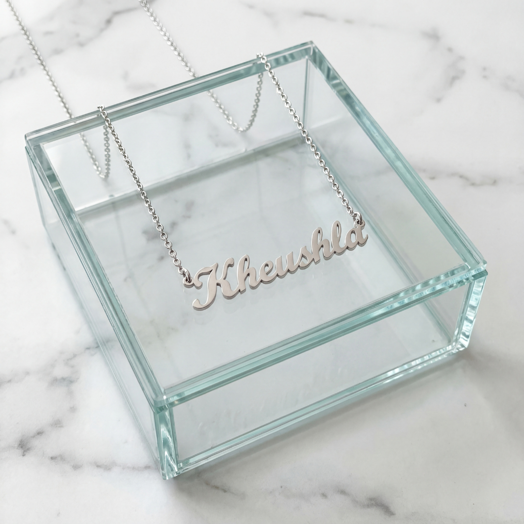 Silver cursive personalized name necklace spelling Kheushla displayed on a glass jewelry box on white marble