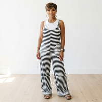 A woman modeling a sleeveless black and white striped jumpsuit with a white tank top underneath, standing in a bright room with light wood floors and a white wall.