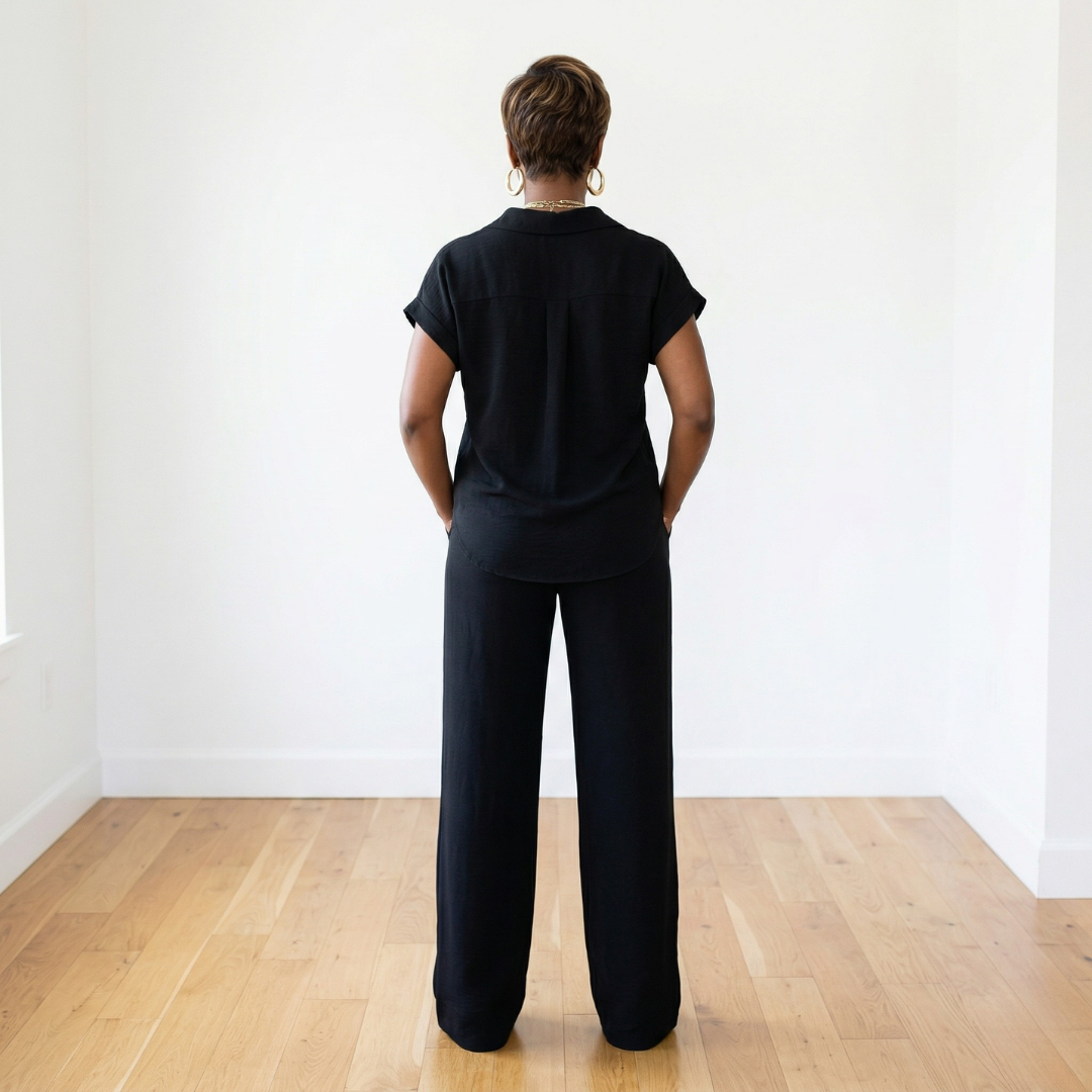 African American woman wearing a black two-piece shirt and pants set with a button-down top and long wide-leg pants in a white room with hardwood floors