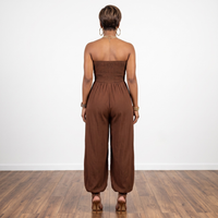 A woman viewed from behind wearing a brown strapless jumpsuit with a smocked bodice and wide-leg pants, standing on a wood floor against a white wall.