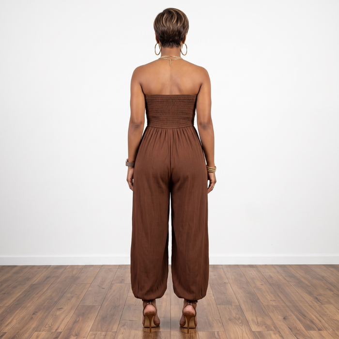 A woman viewed from behind wearing a brown strapless jumpsuit with a smocked bodice and wide-leg pants, standing on a wood floor against a white wall.