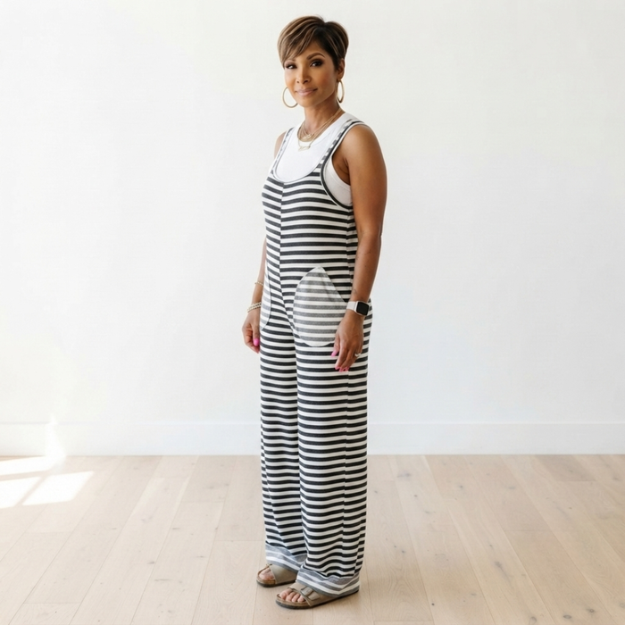 A woman modeling a sleeveless black and white horizontally striped jumpsuit with a white undershirt, standing on a light wood floor against a white wall.