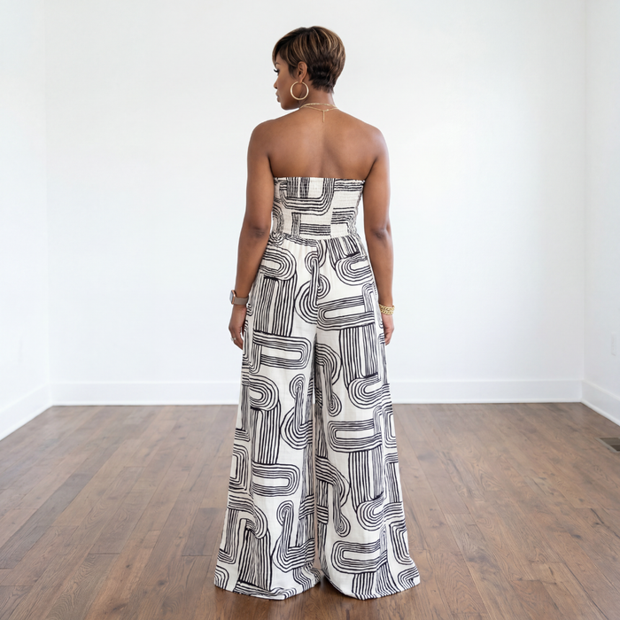 Woman wearing a strapless, patterned jumpsuit standing on a wooden floor with a white wall background.
