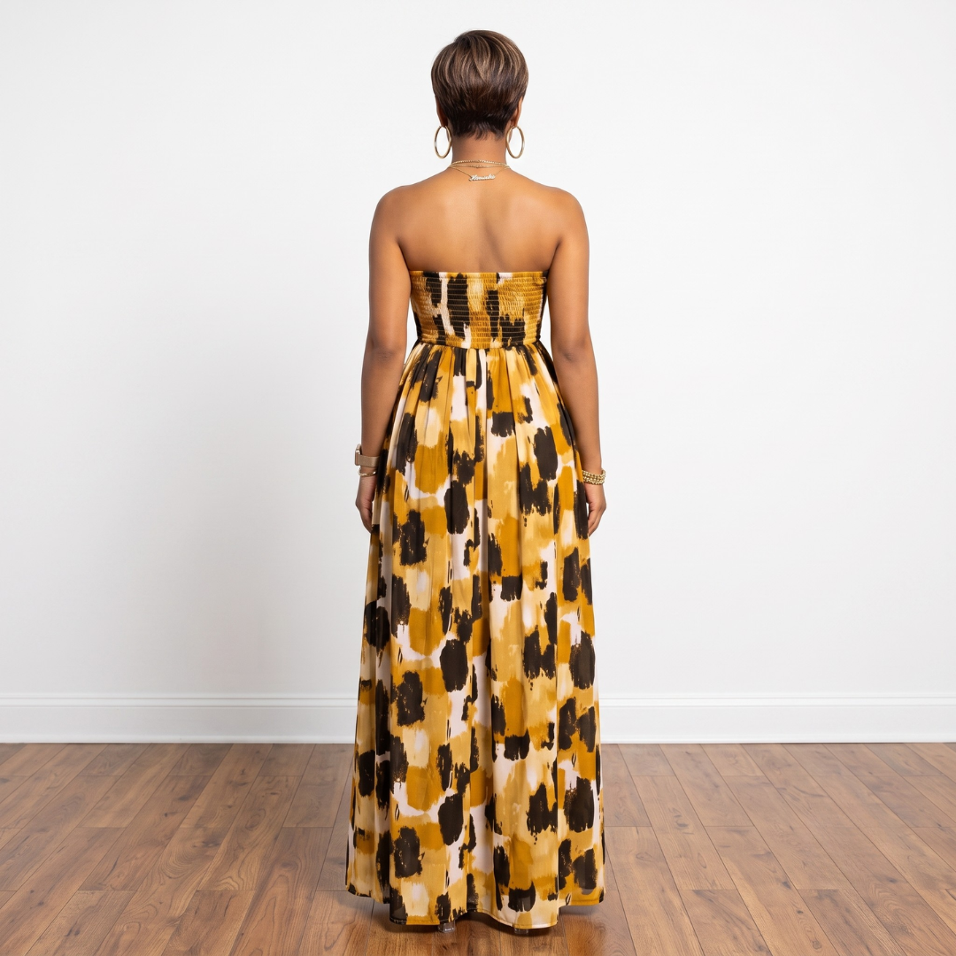 A woman stands with her back to the camera wearing a strapless maxi dress with a mustard yellow, black, and cream abstract pattern. The photo is taken in a minimalist room with a white wall and wooden floor.