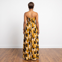 A woman stands with her back to the camera wearing a strapless maxi dress with a mustard yellow, black, and cream abstract pattern. The photo is taken in a minimalist room with a white wall and wooden floor.