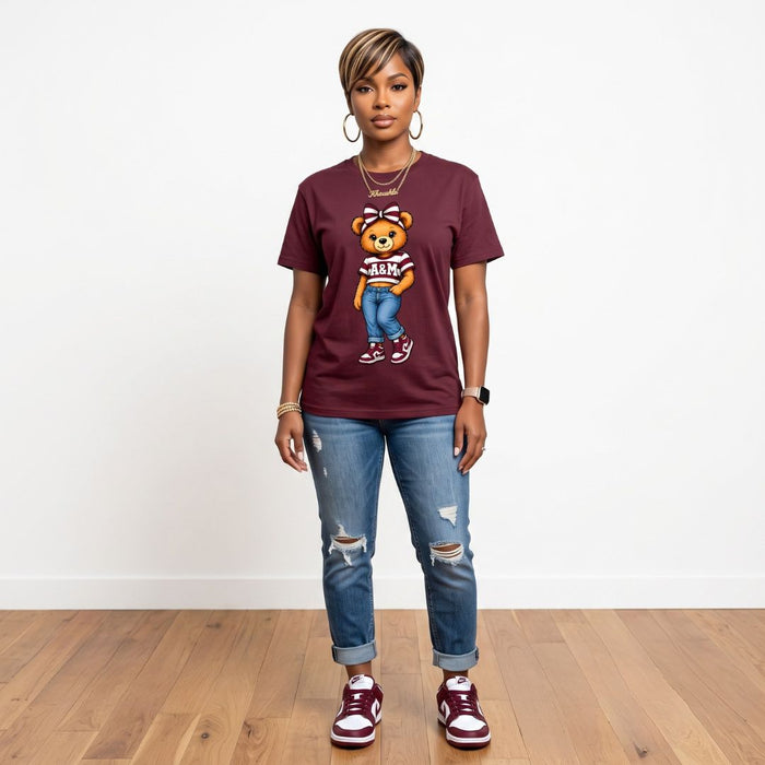 A woman modeling a maroon graphic t-shirt with a cartoon teddy bear wearing 'AIR FORCE' on the front, paired with distressed blue jeans and maroon sneakers, standing against a white wall and wood floor.