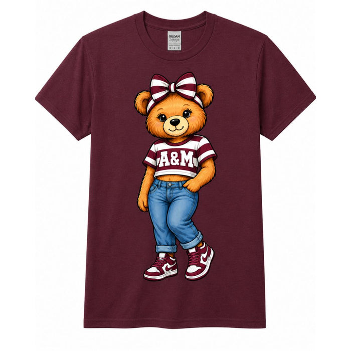 A maroon short-sleeve crewneck t-shirt featuring a cartoon graphic of a brown teddy bear wearing a maroon and white striped A&M shirt, blue jeans, and maroon sneakers.