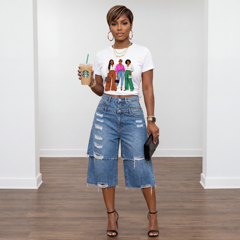 Woman holding a Starbucks cup, wearing a white t-shirt with graphic design and denim shorts, standing against a white wall.